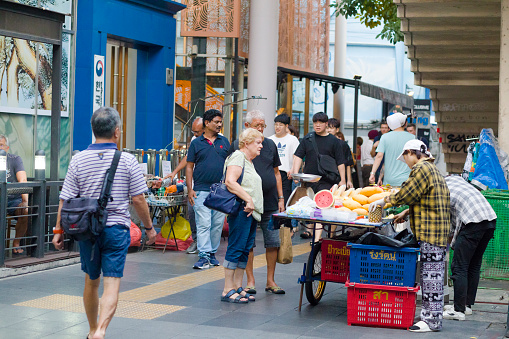 White senior tourist couple is standing st street fruit food market stall on Sukhumvit Road. Couple is wearing  casual clothing. Woman is standing in foreground. Vendor is cutting a pineapple. In foreground and background people are walking on sidewalk. In left background is a small market stall for bags.