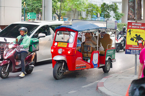 Modern EV tuktuk taxi on Sukhumvitg Rd in Bangkok captured in late afternoon dauly traffic jam. At left side is a delivery express motorcycle driver. Woman and passenger in taxi is wearing a protective face mask and is using mobile phone. In background are queued cars