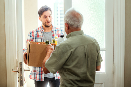 A young adult male volunteers by delivering a box of fresh groceries to an appreciative senior man at his home.