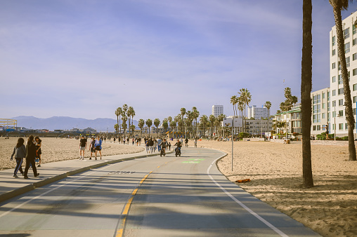 10 March 2024, Los Angeles, USA. Venice beach on a bright sunny day. Crowds of people tourists walking path along pedestrian zone of broadwalk. Cyclists ride on bicycle roadway. Beautiful oceanfront