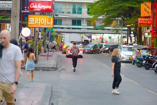 Thai man on EV scooter in Sukhumvit Rd Soi 11 is oassing between people and traffic. Man is wearing a cycling helmet and has a backpack. In background are some cars. People are walking along sidewalk at left side. A woman is crossing street in foreground. In street are bars and restaurants and in background is a small supermarket brand Seven-Eleven