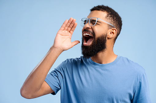 Attractive African American man wearing stylish eyeglasses shouting something, looking away, standing isolated on blue background. Protest concept