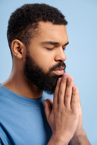 Handsome Christian African American bearded man praying with eyes closed, believing, hoping isolated on blue background, close up. Faith concept, religion