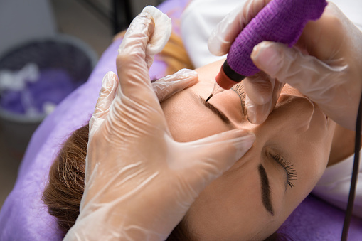 Beauty master performs permanent eyebrow makeup in a beauty salon, close-up. Hands of a cosmetologist doing microblading of eyebrows. Tattoo on the face.