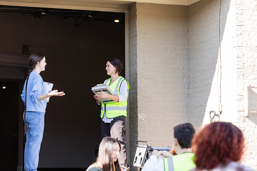 The two female leaders use their clipboards and stand on the loading dock at the volunteer event to help organize people into stations.