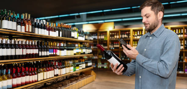 istock Man in casual clothes customer in liquor store scans e-label on wine bottle using his smartphone. 2102233502