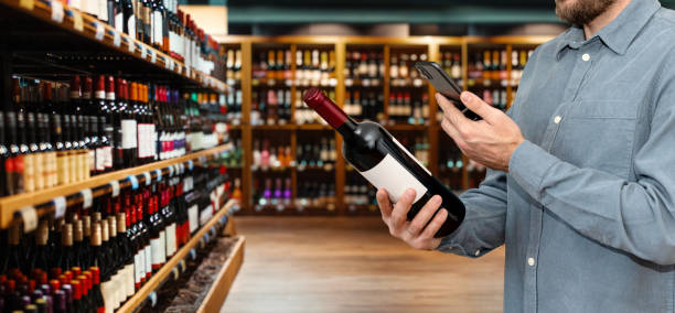 istock Man in casual clothes customer in liquor store scans e-label on wine bottle using his smartphone. 2102235567