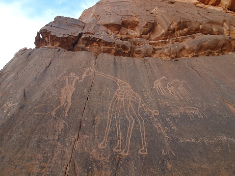 Representing a man carrying a spear, hand outstretched towards a giraffe, giraffes and a horned animal,signs and symbols with a blue sky.