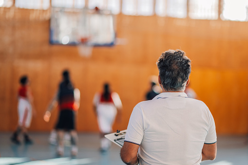 Back view of a senior trainer standing on court indoors and watching basketball training.Rear view of a professional senior basketball coach watching basketball players on training.Basketball training