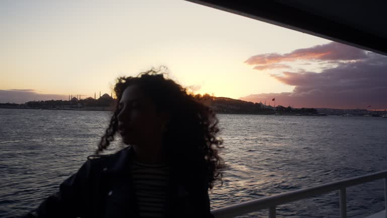 The young woman with curly hair is standing thoughtfully on the ferry, contemplating the impressive Istanbul skyline at sunset.