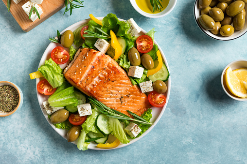 Grilled salmon fish fillet and fresh vegetable salad with feta cheese. Mediterranean food - greek salad and roasted salmon steak on blue background. Barbecue meal.