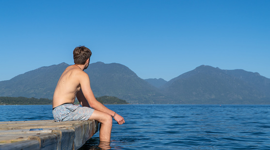 Teenage boy in swimwear sitting on the lake dock, gazing at the horizon with mountains in the background. Perfect summer snapshot.