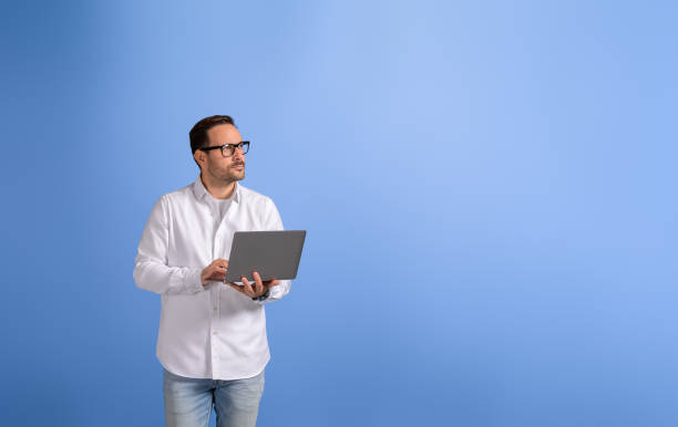 istock Thoughtful male professional with wireless computer looking away and standing over blue background 2102422899