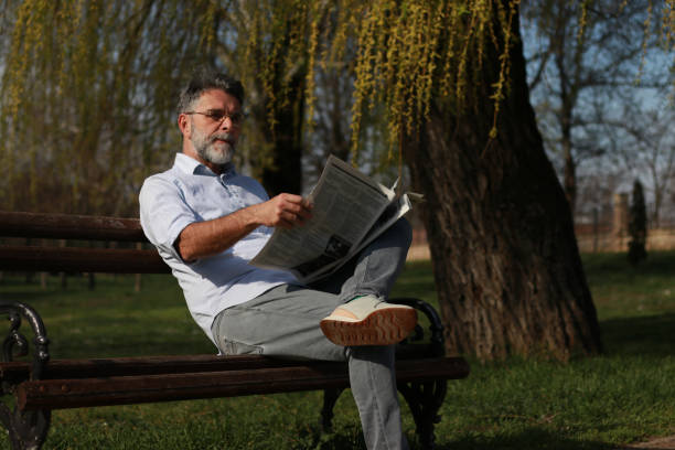 istock Older bearded man reading newspaper in public park 2102440727