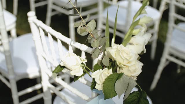 Close up view of Wedding floral decorations of flowers in pastel faded colors slow motion.White wooden chairs decorated with flowers. Frame for outside wedding ceremony in park.