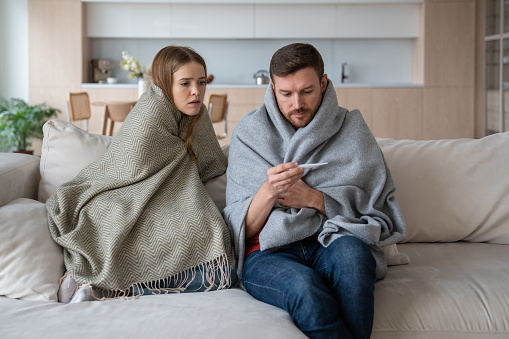 Sick couple sitting on sofa wrapped in blanket, checking temperature on thermometer. Wife and husband with symptoms of respiratory virus, influenza, fever, in need of medical, treatment and isolation
