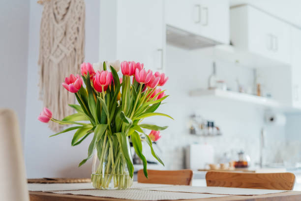 Stylish and modern boho, scandi interior of open space white kitchen with pink tulip flowers in vase on the wooden kitchen counter table, macrame on the wall. Cozy Design home decor. Selective focus