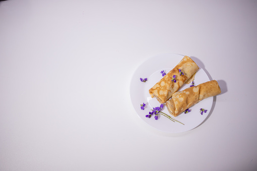 Homemade  European thin  pancakes rolled up on a white plate Delicious French pancakes rolled up  decorated with purple violets on a  white plate with white background