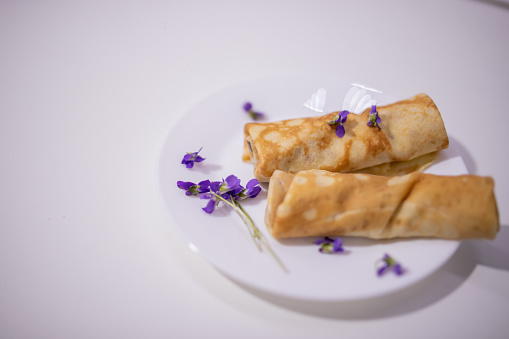 Homemade  European thin  pancakes rolled up on a white plate Delicious French pancakes rolled up  decorated with purple violets on a  white plate with white background