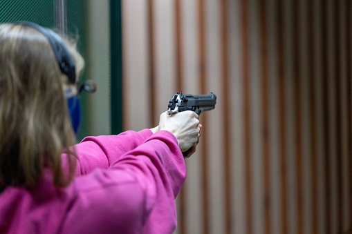 Teenager in a pink tracksuit wearing noise-cancelling headphones aiming a 9 mm gun at a shooting range