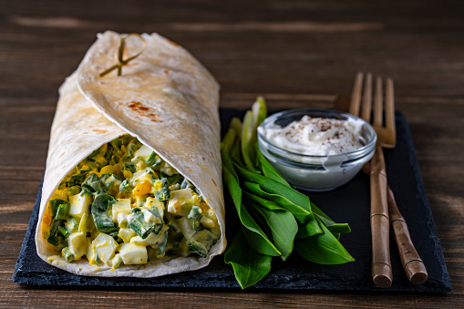Homemade burrito wraps with boiled eggs, green wild garlic and sour cream for healthy breakfast on wooden board, close up