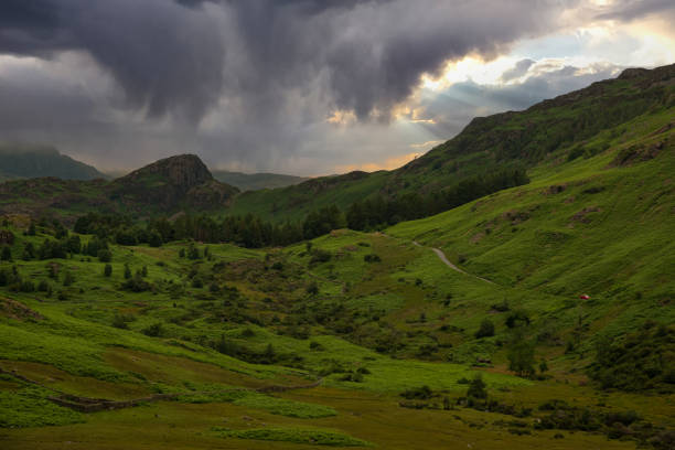 AERIAL: Threatening rain clouds hover above rolling green hills of Lake District, hinting at an upcoming downpour in a peaceful natural environment. Moody weather in a picturesque mountain region.