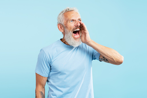 Portrait of mature angry bearded man shouting announcing something, looking away wearing stylish casual blue t shirt standing isolated on blue background. Communication concept