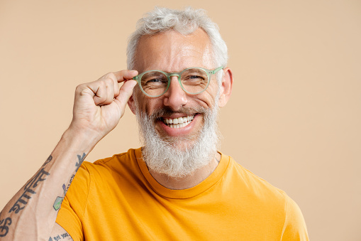 Smiling successful mature man, wearing stylish eyeglasses and casual yellow t shirt, looking at camera, standing isolated on beige background. Vision concept