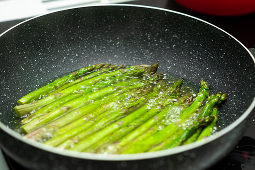 A pan of green asparagus is cooking in a pan. The asparagus are in a stir fry and are being cooked in a pan with oil