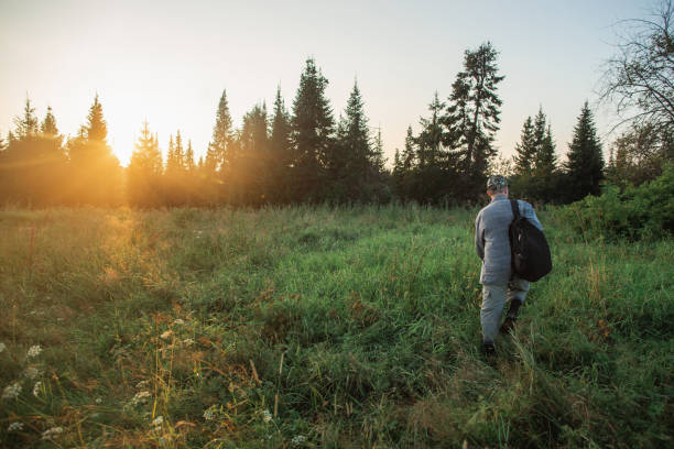 istock Man with backpack stands in field at sunset, surrounded by trees and grassland 2102717031