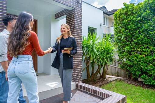 Real estate agent showing a couple a new house. The house is contemporary. All are happy and smiling and shaking hands. The couple are casually dressed and the agent is in a suit. The house has a brick facade