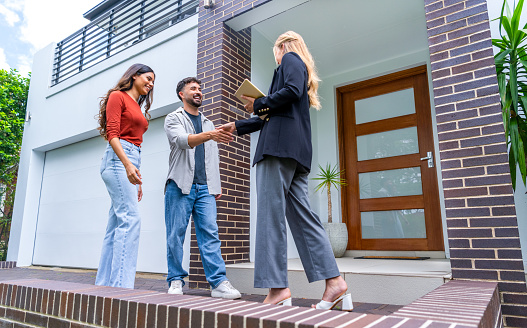 Real estate agent showing a couple a new house. The house is contemporary. All are happy and smiling and shaking hands. The couple are casually dressed and the agent is in a suit. The house has a brick facade