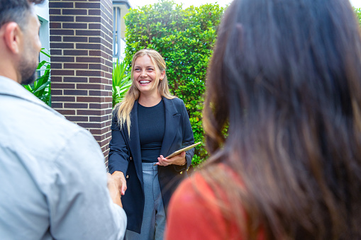 Real estate agent showing a couple a new house. Some of the front garden can be seen. All are happy and smiling and shaking hands. The couple are casually dressed and the agent is in a suit. The house has a brick facade