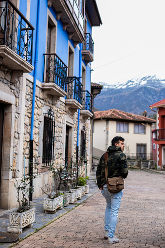 Peruvian man in camo coat, brown sling bag, explores vibrant, stone rural houses. Snowy mountains.