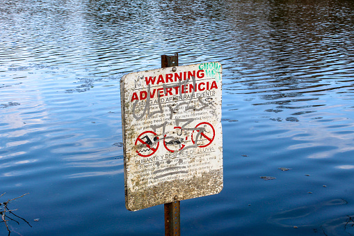 A defaced warning sign, in both English and Spanish, that prohibits swimming, fishing and wading seen against a lake backdrop