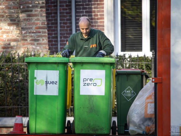 Worker, garbage man, of Suez Prezero recycling handing garbage bins in Maastricht. Prezero is a waste disposal company. Maastricht, Netherlands - November 10, 2022: Picture of a garbage man managing green bins in maastricht, Netherlands. PreZero is a waste management and recycling company. The company's history began with the creation of the PreZero brand by GreenCycle, a company of the Schwarz Group, in 2018 as an online platform for obtaining quotes for recyclable and waste disposal. PreZero has expanded its operations through acquisitions, including the acquisition of the Tönsmeier Group, a German waste disposal and recycling company with nearly a century of experience, and the Suez disposal and recycling locations in various European countries. PreZero operates internationally with locations in Europe and the United States, focusing on developing solutions for recycling and resource conservation across a network of over 430 locations on two continents. The company aims to advance circular economy principles and reduce reliance on finite resources by transforming waste into new raw materials. Headquartered in Germany, PreZero is part of the Schwarz Group, one of Europe's leading retail groups, which also includes supermarket chains like Lidl and Kaufland. The company's activities extend beyond waste management to encompass the entire recycling loop, and it is involved in various sustainability and recycling initiatives both within the Schwarz Group and with external partners public service employee stock pictures, royalty-free photos & images