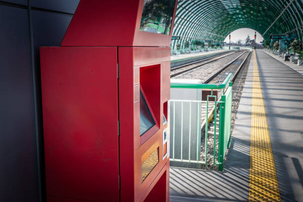 At a bustling train station, a modern, red ticket vending machine stands ready to serve the needs of commuters. It's a typical sight in today's fast-paced world, where efficiency and self-service technology are essential for the on-the-go lifestyle. The image captures this machine with its digital screen off to the side, implying its readiness for travelers to make their selections. In the background, the platform extends into the distance under a contemporary, arched, translucent roof that shelters from the elements, enhancing the sense of a journey about to begin. The foreground focuses on the functionality and solitude of the ticket machine, a silent facilitator of daily commutes and sporadic adventures. It reflects a common yet indispensable aspect of urban infrastructure, symbolizing movement and the pulse of city life. This image, with its juxtaposition of stillness and the potential for motion, is emblematic of the traveler's experience  the moments of quiet anticipation before the hustle of transit. Perfect for projects and pieces discussing urban planning, public transportation, modern conveniences, or the rhythm of city life, this photo encapsulates a shared experience in public spaces, where technology and human paths intersect. It's also a nod to the design and operation of public services, hinting at the network of systems that keep a city humming and connected.