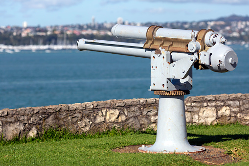 A pair of shiny gold metal or brass binoculars fixed to a tripod and pointed to observe the view of the pacific ocean and blue sky on a sunny day. The binoculars point in the direction of the sea ocean waterfront across the view of blurred grass and a hedge in a garden backyard.