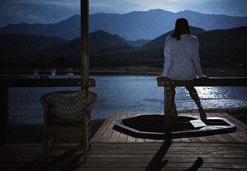 Rear view of a woman looking out at the scenic moonlit landscape while standing on the deck of a log cabin during a vacation in a nature reserve