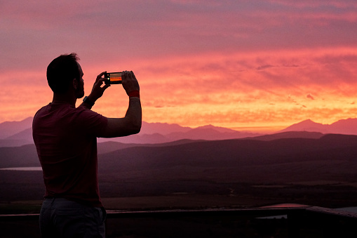 Rear view of man standing on observation deck in nature and taking picture of beautiful sunset sky over the mountains with his mobile phone