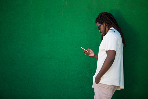 Black man leaning against a green wall checking balance on his cell phone. Wears a white shirt, dread locs, sunglasses
