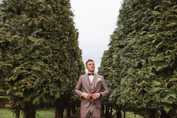 茶色のスーツを着た新郎は、緑の木々を背景にポーズをとり、ジャケットを調節します。結婚式の肖像画。 - fashion model personal accessory suit tying ストックフォトと画像