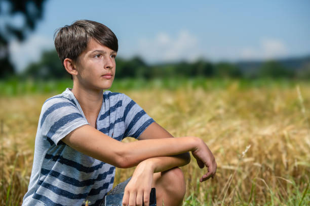 A boy kneels in a field of ripening barley and admires it. A teenager runs his hand over an ear of barley in a field.