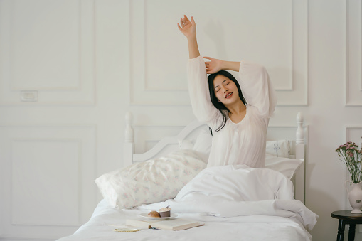 A woman is in bed, stretching and yawning. She is wearing a white shirt and is sitting on a bed with a book and a cupcake on a plate. Concept of relaxation and leisure
