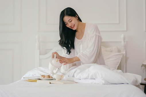 A woman is pouring tea into a cup while sitting on a bed. Concept of relaxation and comfort, as the woman is enjoying a quiet moment in her bedroom