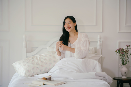 A woman is sitting on a bed with a book and a plate of food in front of her. She is smiling and she is enjoying her time. The bed is covered in white sheets and pillows