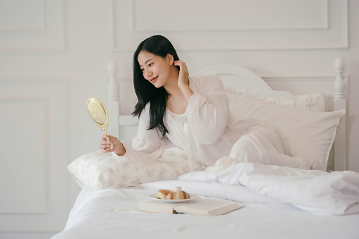 A woman is sitting on a bed with a mirror in front of her. She is wearing a white shirt and is brushing her hair. The bed is covered with white sheets and pillows