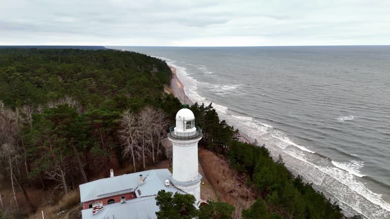 A lighthouse in Uzava is on a rocky Baltic Sea shoreline, large body of water in the background. The lighthouse is tall and white, and it is surrounded by trees. The scene is peaceful and serene