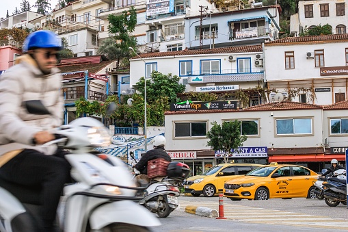 Fethiye, Turkey - December 8, 2022: Man on scooter rides through narrow street in small town with shops and cafes on both sides.