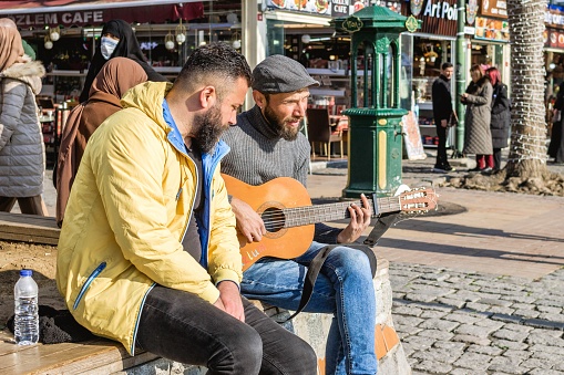 Istanbul, Turkey - December 29, 2022: Two men are sitting on bench by the street and one of them playing guitar.